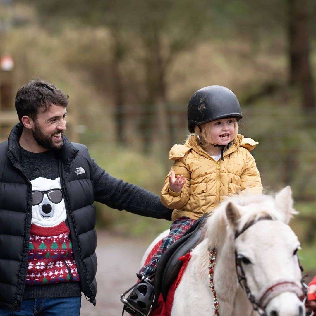 Child wearing a helmet rides a small white pony, smiling; an adult steadies them while walking beside. Outdoor wooded path. Visible text: "The North Face" on the adult’s jacket.