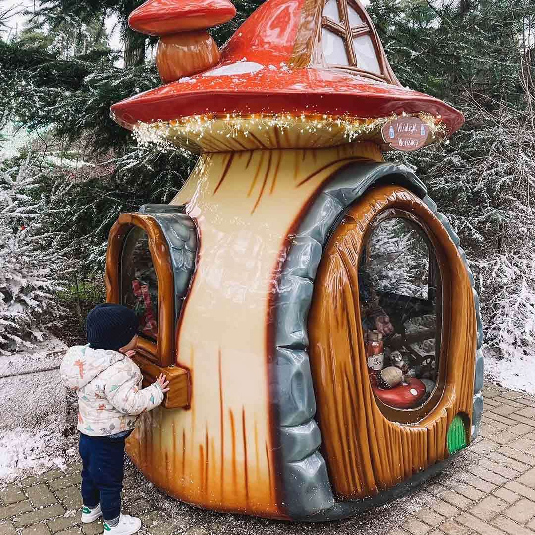 Child touches a window, peering into a mushroom-shaped miniature house. Fairy lights trim the roof amid snowy evergreen trees and a paved path. Text on sign: “Wishlight Workshop”.