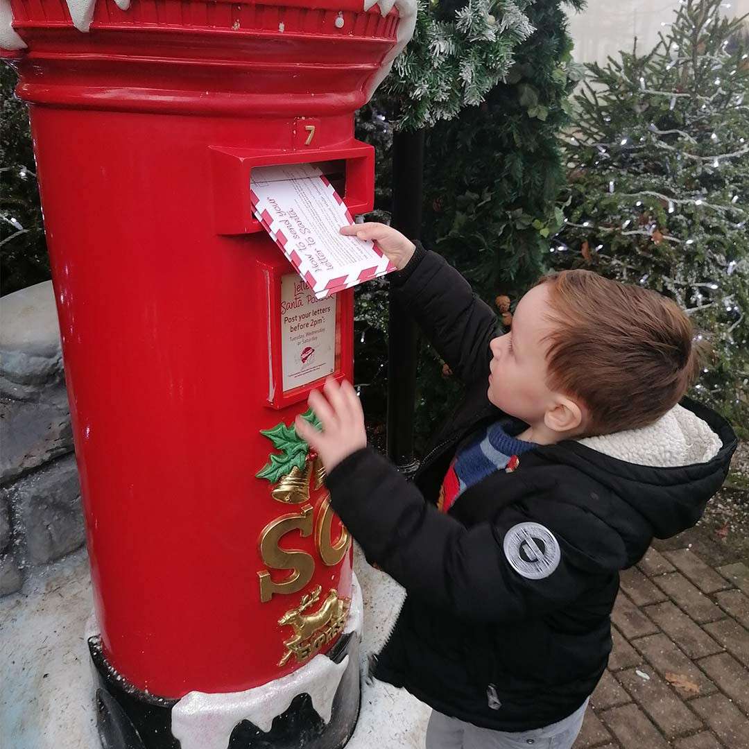 Child inserts letters into a large red Santa postbox amid snowy Christmas decorations and fir trees. Text: 7; Letters to Santa; Post your letters Before 2pm.