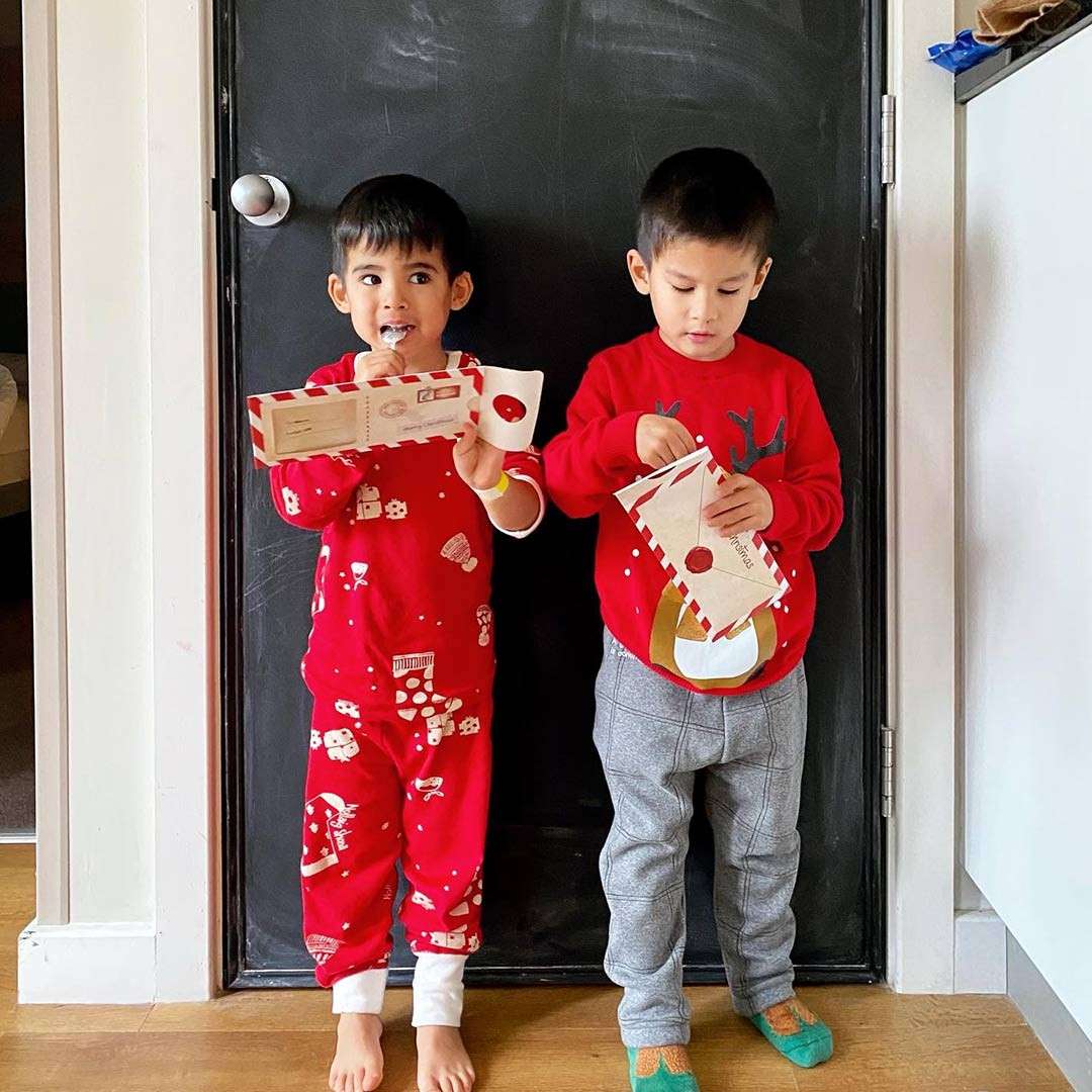 Two children open red-and-white envelopes; one licks a treat while the other examines contents, standing barefoot in festive red pajamas and sweater against a black door inside a home.