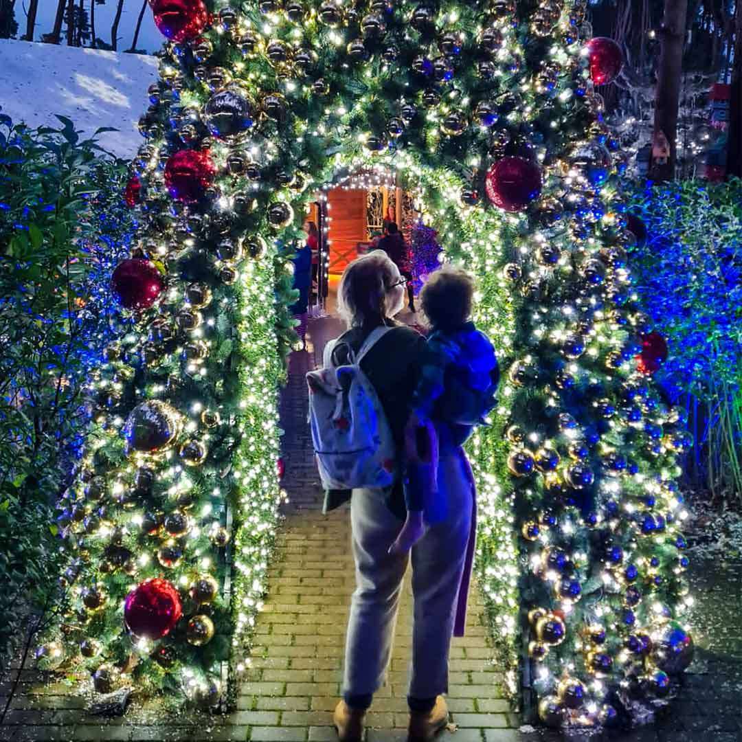 An adult carrying a child gazes up while standing beneath a brightly lit evergreen archway, decorated with red and gold ornaments, along a brick path in a nighttime festive garden.
