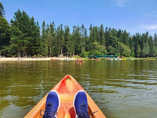 Kayak bow with blue-sneakered feet points forward, drifting on a calm lake; across the water, a sandy beach, scattered kayaks, and dense evergreen forest under a clear blue sky.