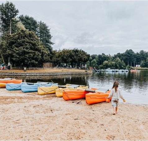 Kayaks rest in a row; a child walks toward them on a sandy shore, amid a calm lake, tree-lined banks, and distant docks under an overcast sky.