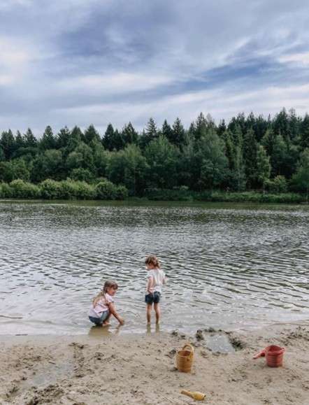 Two children play in shallow lake water, one squatting and splashing, another standing. Sandy beach with buckets and shovel in foreground; calm water and dense forest under cloudy sky.