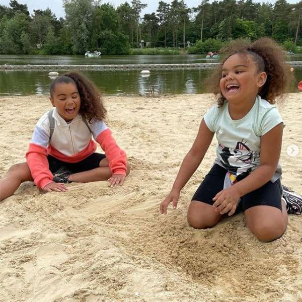 Two children laugh while digging in sand, sitting cross-legged and kneeling. Context: a sandy lakeshore with calm water, floating buoys, and tree-lined background under overcast sky.
