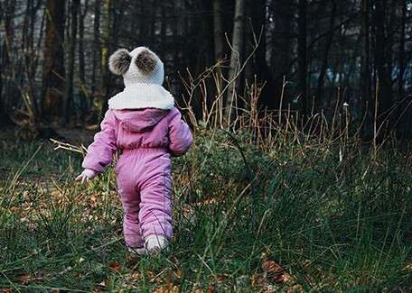 Child in a pink snowsuit walks away across a leafy, grassy forest floor, wearing a white hat with two pom-poms, moving toward dense trees and underbrush in dim, wintry light.