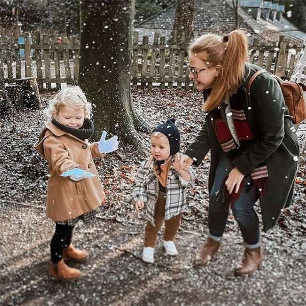 Adult with backpack kneels, holding a toddler’s hand; nearby, another child extends gloved palm catching falling snow; context: wooded park path with trees, leaf-strewn ground, and wooden fence in background.