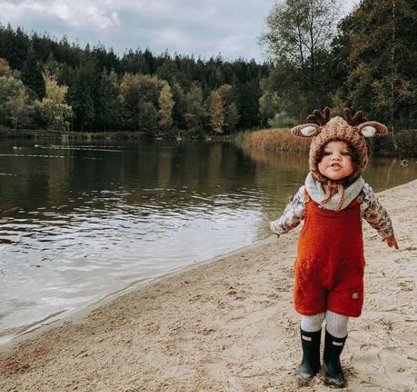 Child in red overalls and reindeer hat stands smiling, arms out, wearing rain boots, on a sandy lakeshore; calm water and autumn trees under a cloudy sky behind.
