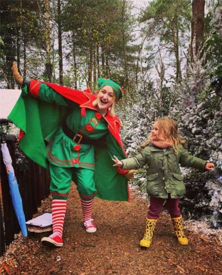 Elf-costumed performer, in red-and-green outfit and striped socks, balances with arms spread; a child in coat and yellow boots imitates, smiling, on a snowy woodland path beside frosted trees.