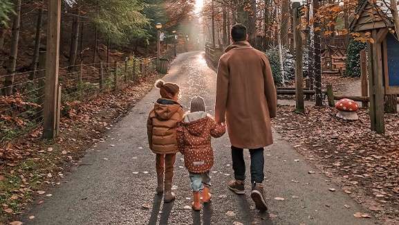 Three people—an adult and two children—walk hand in hand away from the camera along a paved path. Autumn leaves, wooden fences, and forest cabins surround them, with sunlight streaming ahead.