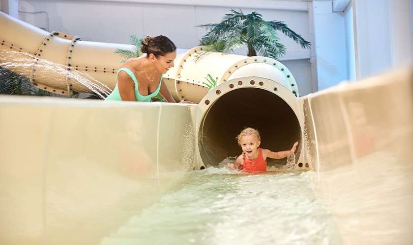 Child slides out of a tube waterslide, splashing into shallow water, while an adult leans over the side watching. Context: indoor water park with plastic slide, spraying water, artificial palm trees.