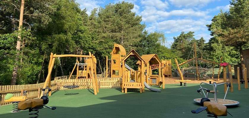 Wooden playground structures stand ready: slides, swings, rope bridge, spring riders, and climbing frames. Set on green safety surface, fenced areas, surrounded by tall trees under a partly cloudy sky.