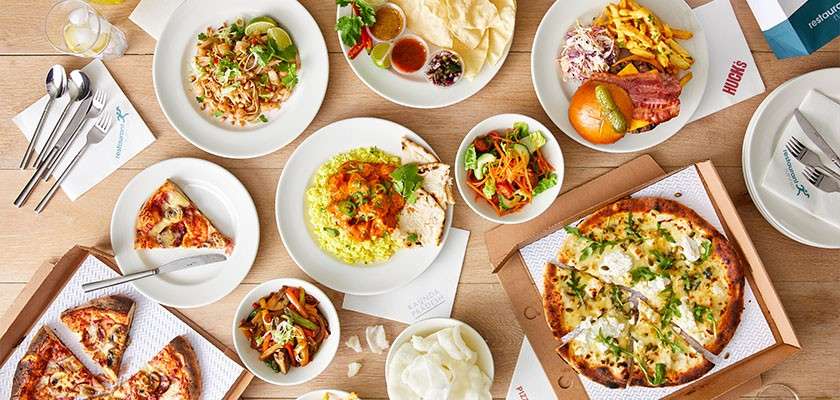 Assorted dishes sit arranged on a wooden table: pizzas, pasta, salads, a burger with fries, sauces, and drinks, surrounded by menus, boxes, and cutlery. Visible text: "HUGO'S".