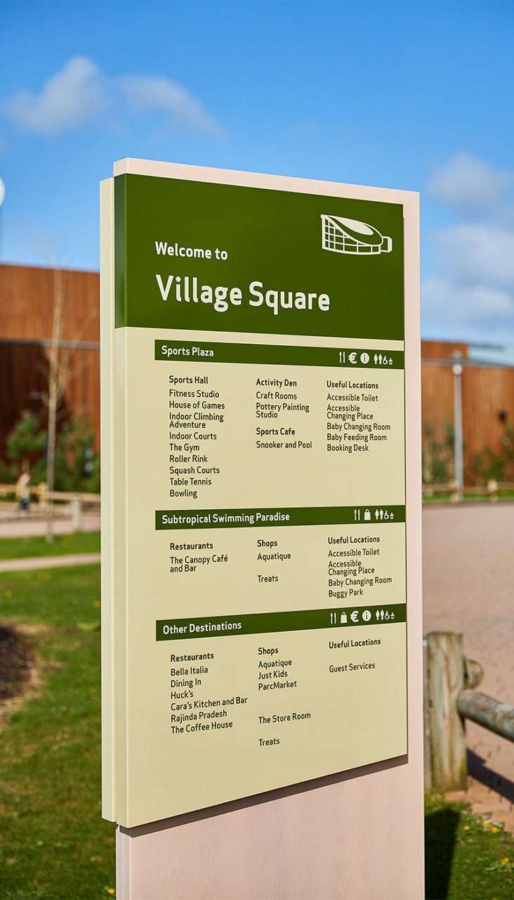Wayfinding sign lists attractions and facilities, standing beside a path in a landscaped area with buildings.

Text:
Welcome to
Village Square

Sports Plaza
- Sports Hall
- Fitness Studio
- House of Games
- Indoor Climbing Adventure
- Indoor Courts
- The Gym
- Roller Rink
- Squash Courts
- Table Tennis
- Bowling
- Activity Den
- Craft Rooms
- Pottery Painting Studio
- Sports Cafe
- Snooker and Pool
Useful Locations
- Accessible Toilet
- Accessible Changing Place
- Baby Changing Room
- Baby Feeding Room
- Booking Desk

Subtropical Swimming Paradise
Restaurants
- The Canopy Cafe and Bar
Shops
- Aquatique
- Treats
Useful Locations
- Accessible Toilet
- Accessible Changing Place
- Baby Changing Room
- Buggy Park

Other Destinations
Restaurants
- Bella Italia
- Dining In
- Huck’s
- Cara’s Kitchen and Bar
- Rajinda Pradesh
- The Coffee House
Shops
- Aquatique
- Just Kids
- ParcMarket
- The Store Room
- Treats
Useful Locations
- Guest Services