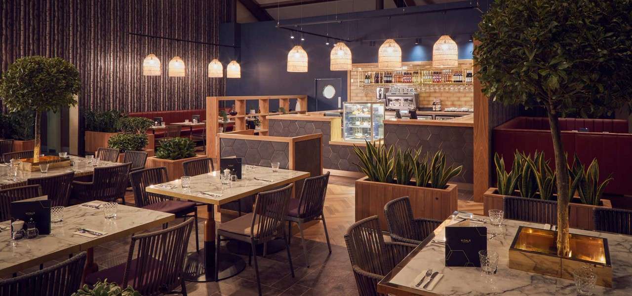 Dining tables set with menus, glasses, and cutlery, while a counter displays cakes. Surrounding a modern restaurant with wicker pendant lights, wood partitions, potted plants, marble tops, and red booths.