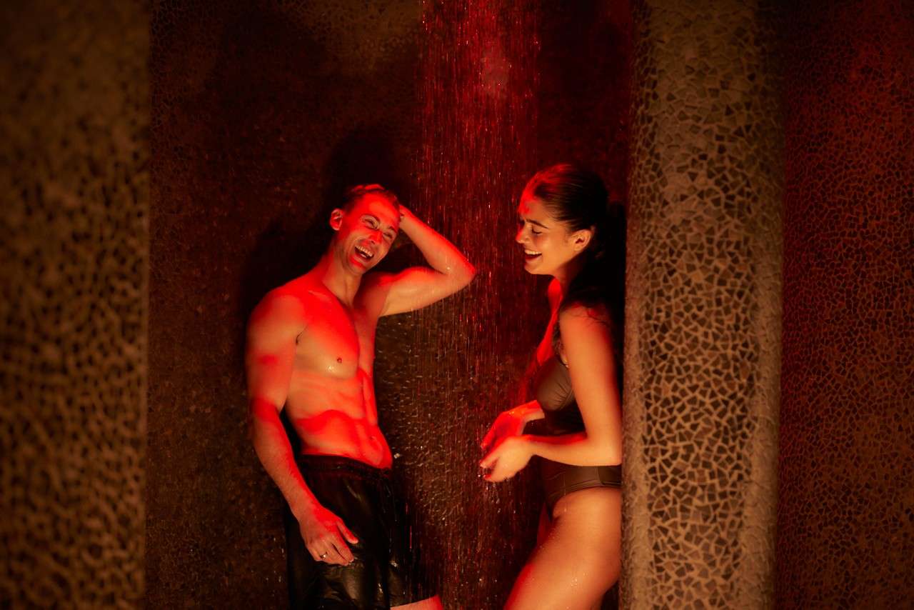 Two adults in swimwear laugh while standing under a red-lit shower, water cascading over them, in a dim, mosaic-tiled spa alcove.