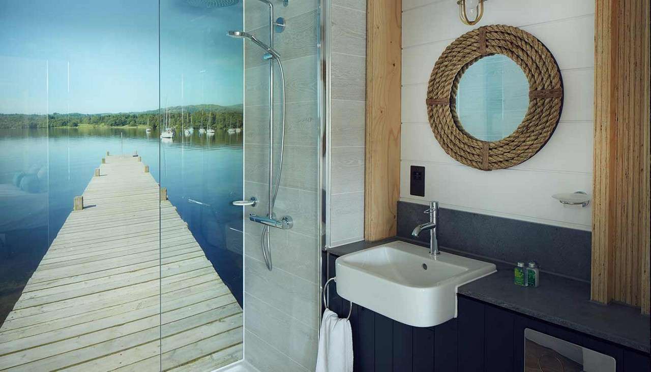 Shower enclosure displays a mural of a wooden dock extending into a calm lake. Beside it, a modern sink and rope-framed round mirror above a gray counter with towel ring.