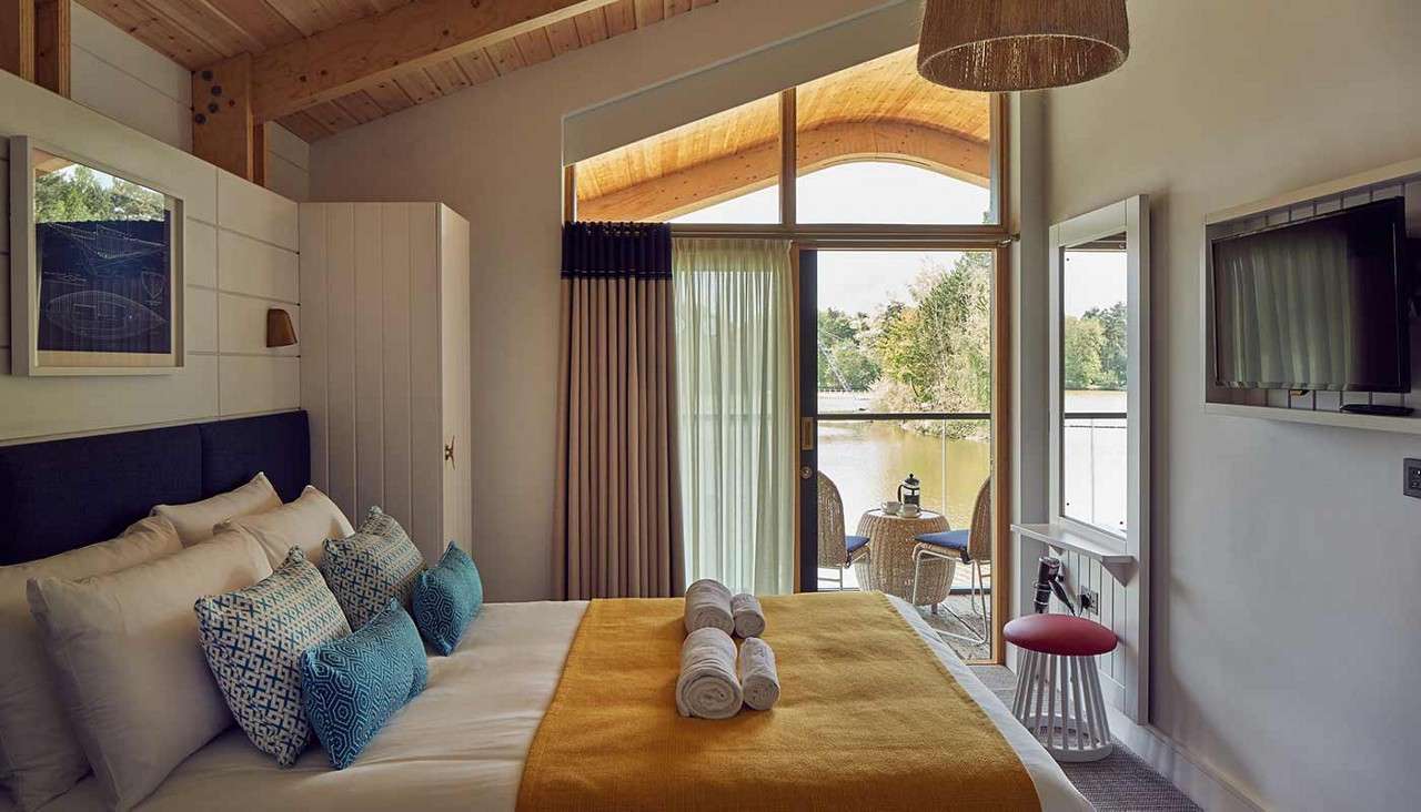 Bed displays folded towels and patterned pillows, facing sliding glass doors; beyond, two wicker chairs flank a small table on a balcony overlooking a lake, beneath a wooden vaulted ceiling.