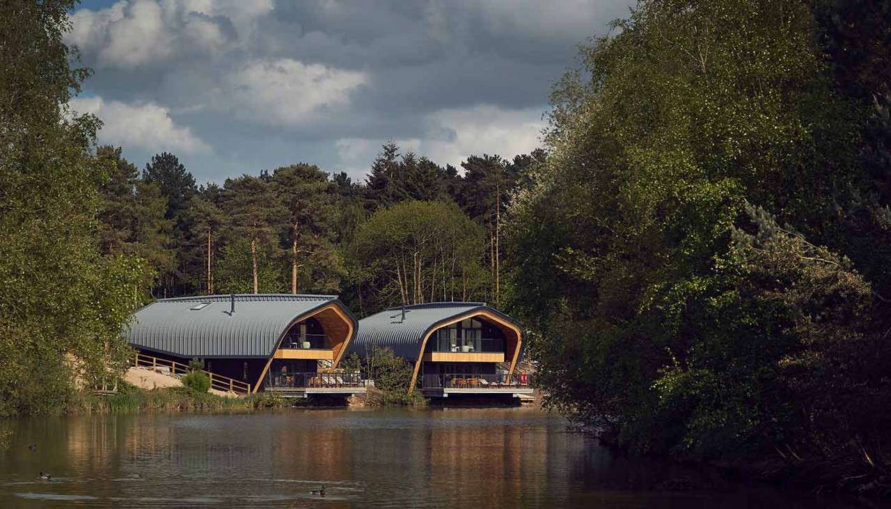 Two modern, curved-roof lakeside cabins sit on stilts above calm water; surrounding pine forest and trees frame them under cloudy skies, with ducks swimming in the foreground.