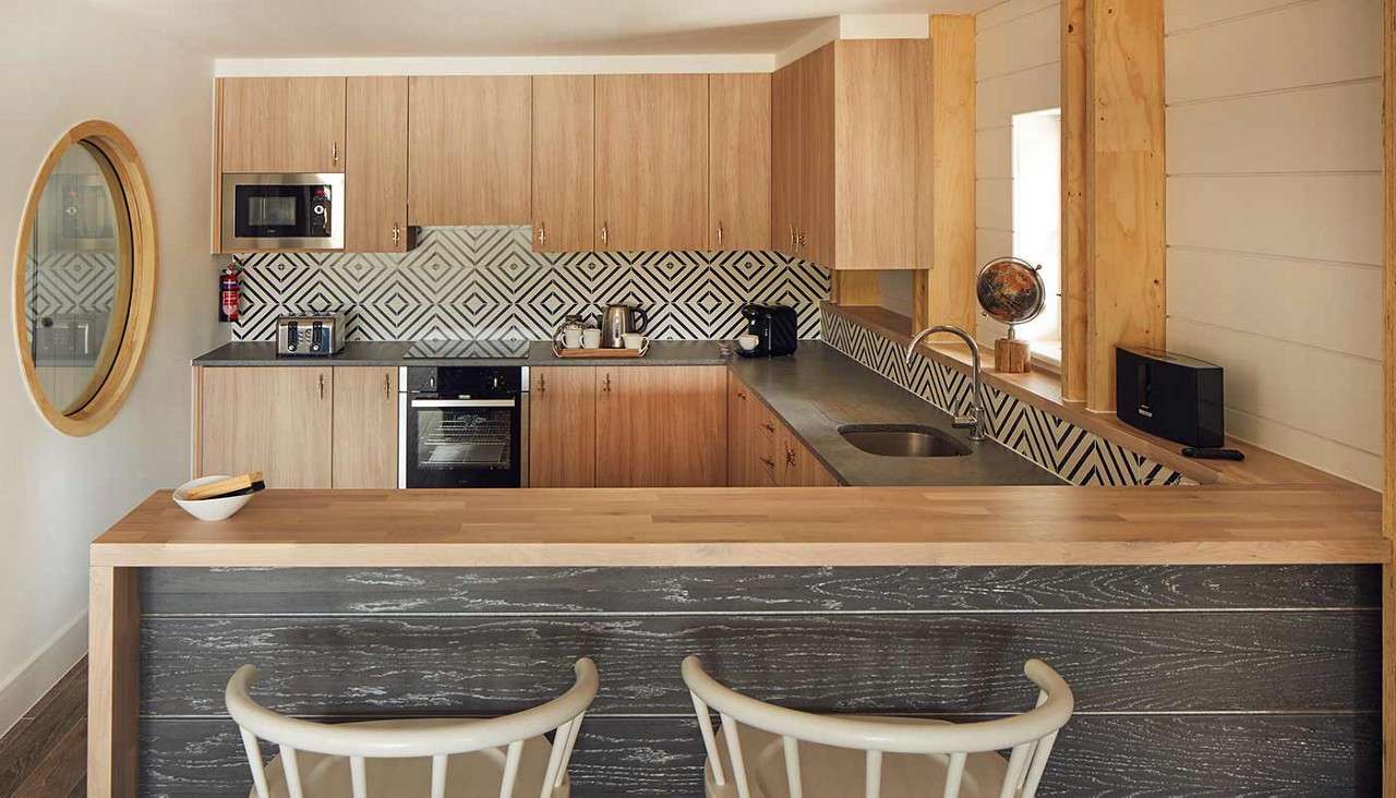 Kitchen showcases light-wood cabinets and island, displaying appliances neatly arranged; oven centered, sink on right, geometric black-and-white backsplash; two stools fronting a bar in a compact, modern interior.