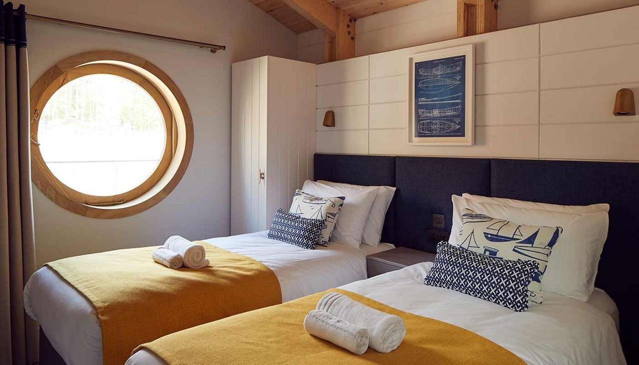 Twin beds rest side-by-side, topped with yellow throws and rolled towels; pillows show nautical prints. Context: wood-accented room with round porthole window, wall lamps, wardrobe, and a framed boat diagram.