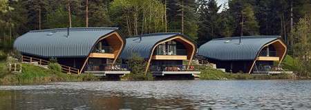 Three modern, curved-roof cabins face the water, their glass-fronted balconies overlooking a calm lake; surrounded by trees and greenery, they sit on stilts near the shoreline in a forested setting.