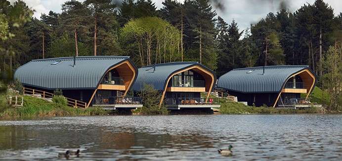 Three curved-roof cabins overlook a lake, their decks facing the water while ducks swim in the foreground; dense conifer forest rises behind, with wooden railings and paths around the lodges.