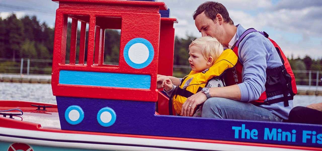 Child wearing a yellow life jacket steers a colorful toy boat while an adult assists, on a calm lake near trees; boat side text reads: "The Mini Fis".