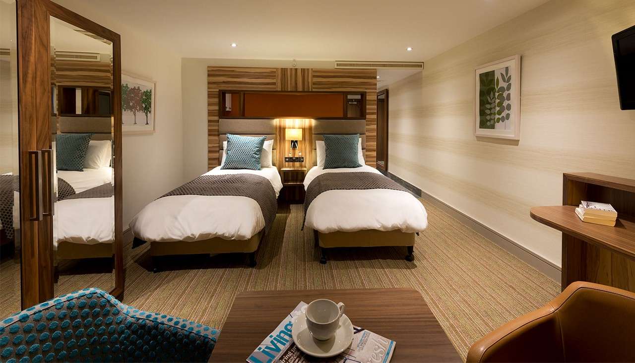 Twin beds stand side by side, lit by a lamp, in a modern hotel room with carpeted floor, wardrobe, and a table holding a cup and magazine titled “Living”.