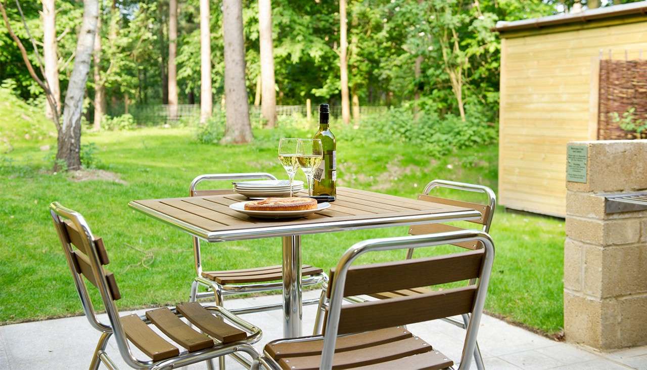 Wooden patio table holds a wine bottle, two filled glasses, and plates with bread; situated on a terrace, bordered by a grassy lawn, tall trees, and a small wooden shed.