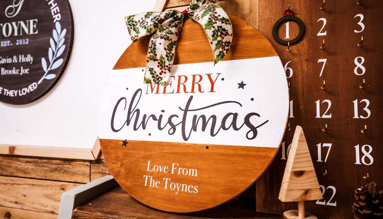 Round wooden Christmas sign leans on a shelf, bow-topped, beside a numbered wooden advent calendar and small wooden tree.

Text:
MERRY
Christmas
Love From The Toynes
EST. 2012
TOYNE
Gavin & Holly
Brooke Joe
1 2 3 12 13 17 18 22
