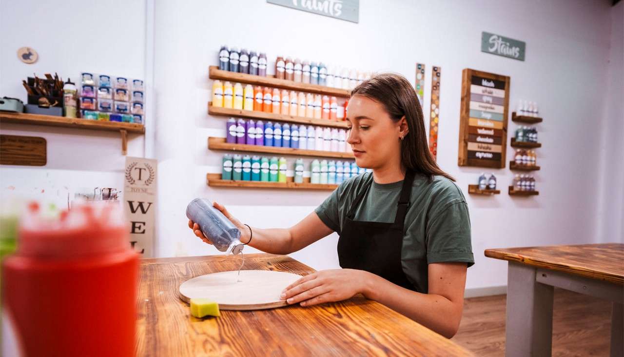 An artist pours liquid from a squeeze bottle onto a round wooden board, in a studio lined with colorful paint bottles and signs reading: Paints; Stains.
