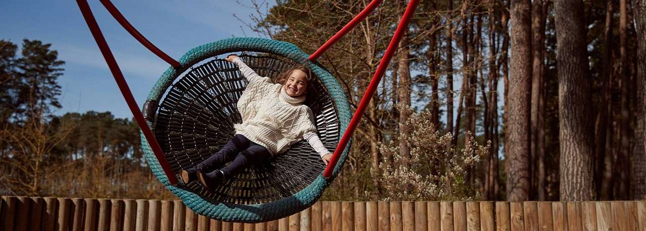 Child reclines and smiles while swinging on a large woven basket swing; red support ropes; background includes wooden fence and tall pine trees in an outdoor playground.