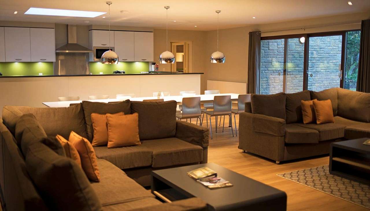 Sectional sofas with orange cushions sit around low coffee tables, facing an open-plan kitchen-dining area with long white table, pendant lights, white cabinets, green backsplash, and sliding glass doors.
