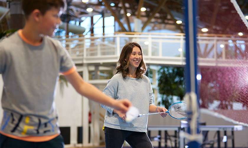 Woman swings a badminton racket, smiling as she returns a shuttlecock. Boy in foreground prepares to hit. Indoor sports center with net, elevated walkway, plants, and table tennis tables.