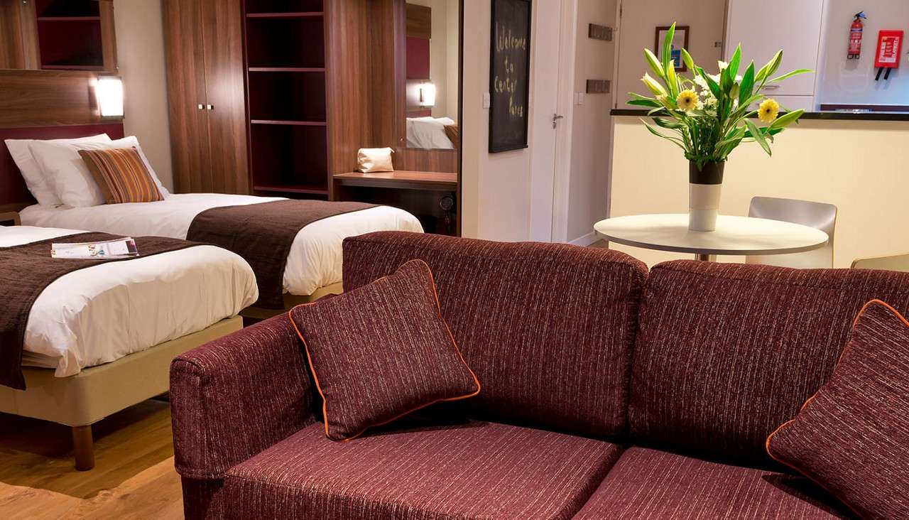 Two single beds rest neatly; a maroon sofa faces a round table holding a flower vase; context: warm hotel-style studio with wooden wardrobe, desk, kitchenette, and fire extinguisher. Text: “Welcome to” (rest unreadable).