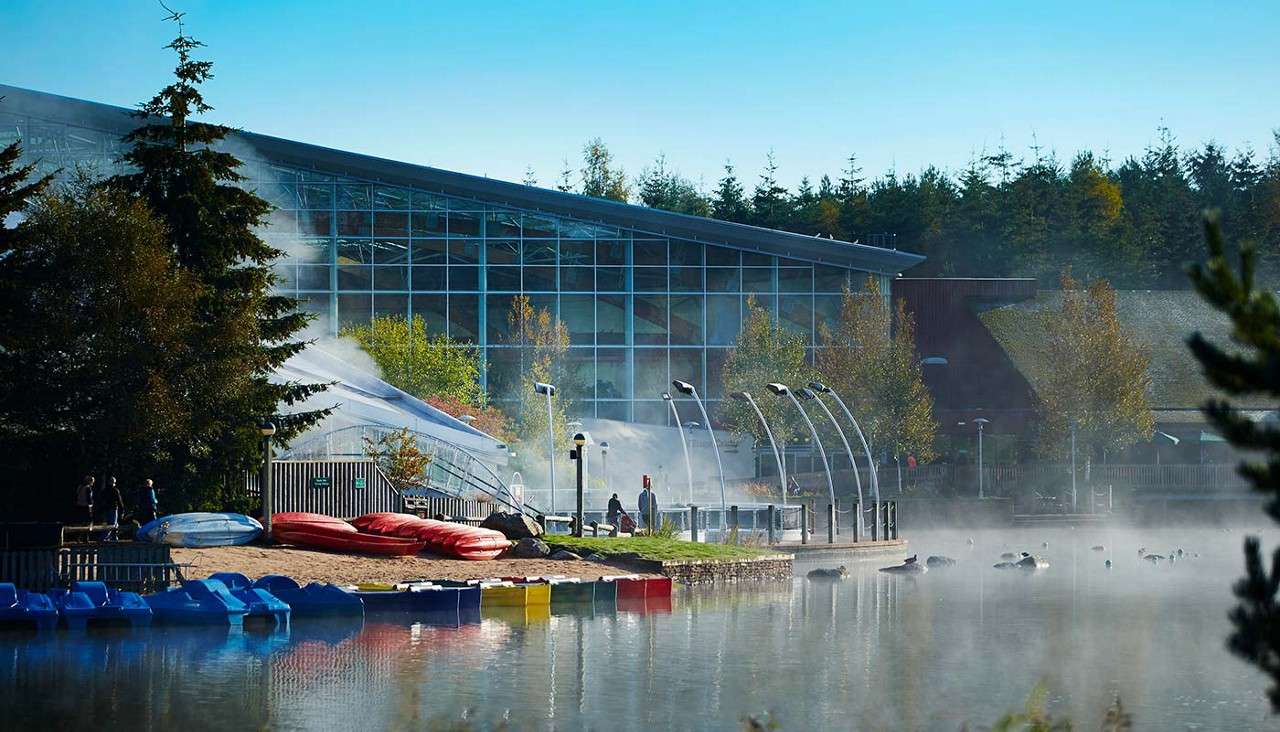 Glass-walled leisure center overlooks a misty lake; kayaks and paddleboats rest on the shore while a few people walk along a curved-canopy waterfront path amid trees.