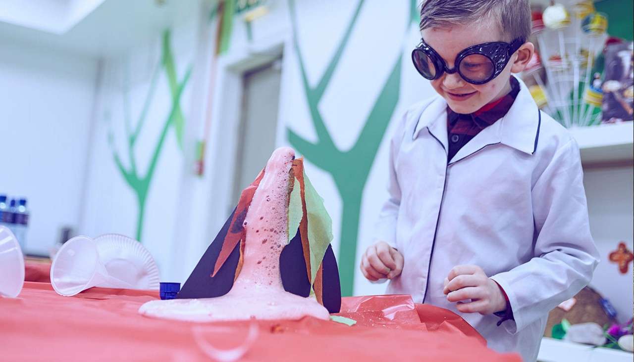 Homemade volcano model erupts pink foamy lava, spilling over paper mountain pieces. A child in goggles and a lab coat watches excitedly at a classroom table with scattered plastic cups.