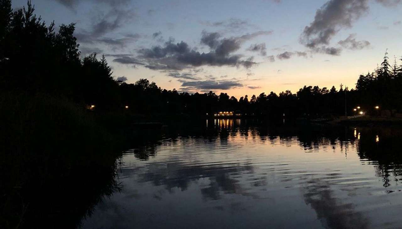 Calm lake water reflects twilight clouds and scattered shoreline lights, rippling gently; silhouetted trees encircle the scene as dusk fades to night beneath a pale orange-blue sky.