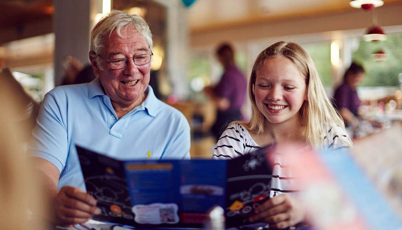 An older man and a girl read menus, smiling, at a casual restaurant table; other diners and warm lights blur in the background.