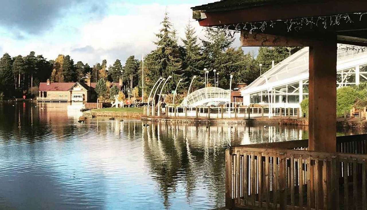 Lakeside boardwalk—person strolling—set within a forested resort. Calm water mirrors pine trees, cabins, and a glass-roofed pavilion; wooden railing and roof frame the foreground.