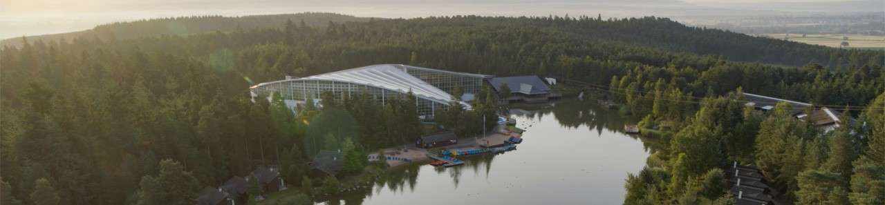 A large glass-roofed resort nestles beside a calm lake, bordered by dense pine forest. Colorful paddleboats line small docks, with scattered cabins and buildings dotting the shoreline under hazy morning light.