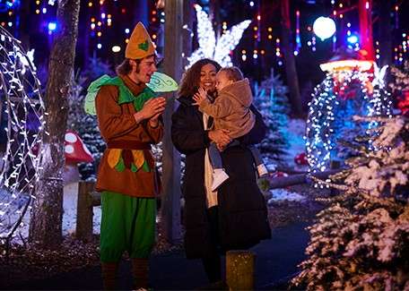 Parent holding a bundled child smiles while a costumed elf claps, amid a winter light display: snow-dusted trees, glowing arches, hanging bulbs, and oversized mushroom decorations along a forest path.