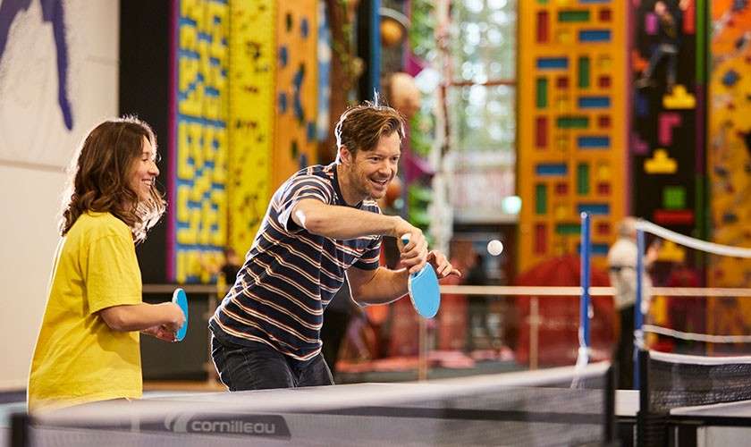 Two adults play table tennis, swinging paddles; an indoor recreation center with colorful climbing walls surrounds them. Visible text: cornilleau.