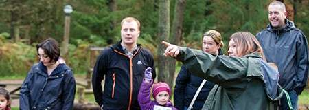 Guide points ahead, instructing a small group of adults and children, who watch attentively, in a forest clearing with trees and simple wooden structures around them.