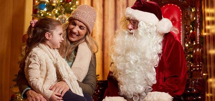 Child smiles and chats with a man dressed as Santa on a red chair. A woman holds the child beside a decorated Christmas tree in a warm wooden room.