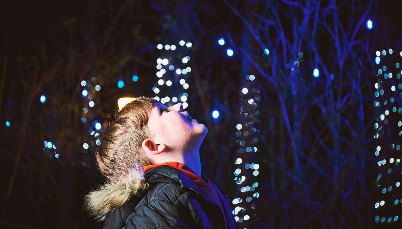 Child in a hooded winter coat tilts head upward, gazing, while standing outdoors at night amid trees decorated with glowing blue and white string lights.