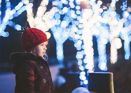 Child in red knit hat gazes sideways, standing bundled in a dark coat; background glows with blue-white holiday lights wrapped around trees at night, softly blurred.