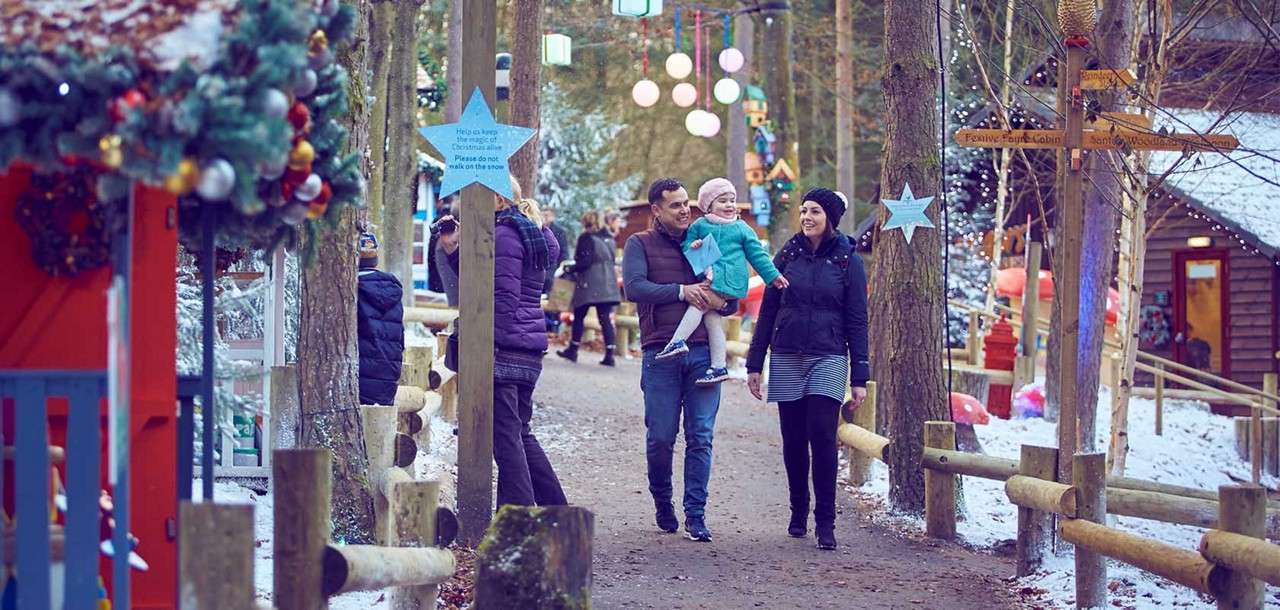 Family—father carrying a toddler, mother walking beside—strolls along a snowy, decorated forest path with lights, cabins, and wooden fences. Visible sign text: “Help us keep the magic of Christmas. Please do not walk on the snow.”