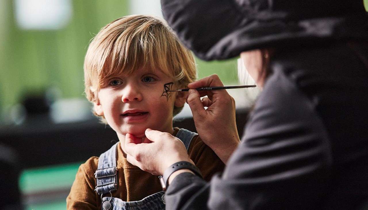 Child sits still while an artist paints a black spiderweb on his cheek, holding his chin with one hand. Indoor setting with soft green background and blurred furnishings.